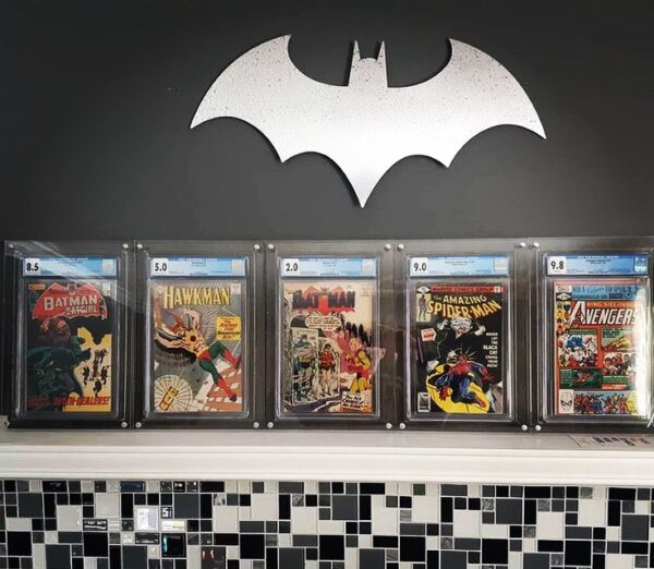 User-generated photo of a room showcasing the Crafti Comics UV-FLEX Display Case with acrylic (clear) backings, a frame designed for graded comic books. This frame features 99% UV-protection with museum-quality acrylic as standard. It can be hung in portrait or landscape orientation using twin engraved mounting holes. Crafti's laser-cut layered designs add depth without applying excessive pressure. The room is painted black and the frames are displayed on a white mantle below a Batman sign.